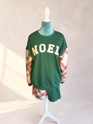 Front view of The Noel Rework, a forest green sweatshirt with cream NOEL lettering, woven holiday textile sleeves, and fringe